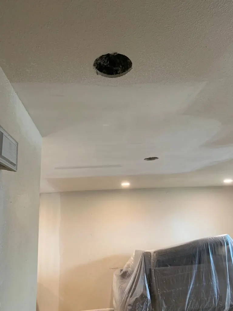 Ceiling drywall repair in progress: an opening in the ceiling with the patch around it skim-coated and furniture covered in plastic